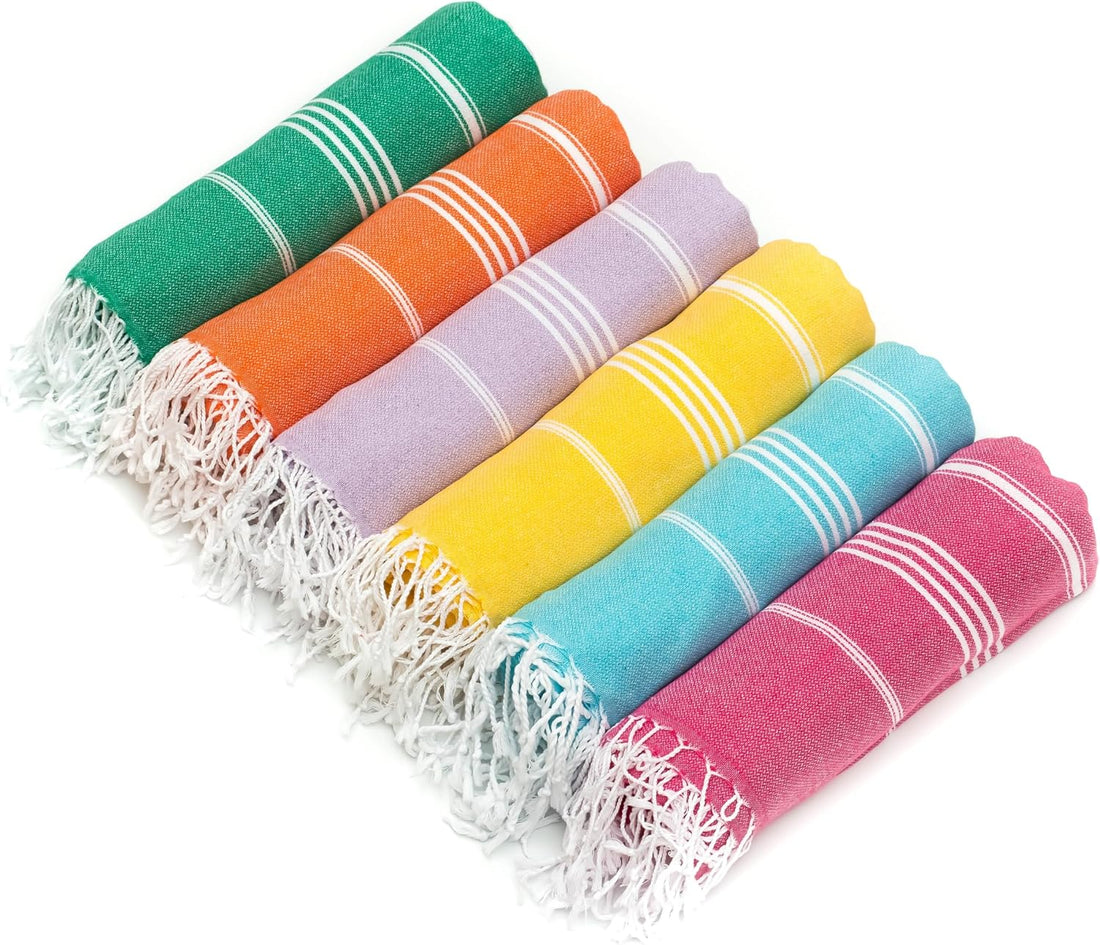 Six rolled Turkish cotton towels fanned out in rainbow colors: green, orange, lavender, yellow, teal, and hot pink. Each towel features white stripe accents and white fringe ends, displayed on a white background.